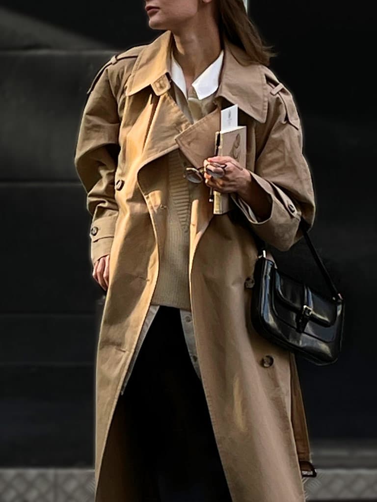 Woman wearing a classic white blouse layered on with a beige sweater and trench coat