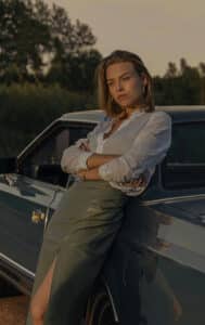A woman wearing a green pencil skirt and white blouse leaning against a car