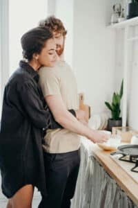 A woman hugging her partner while he is making breakfast.