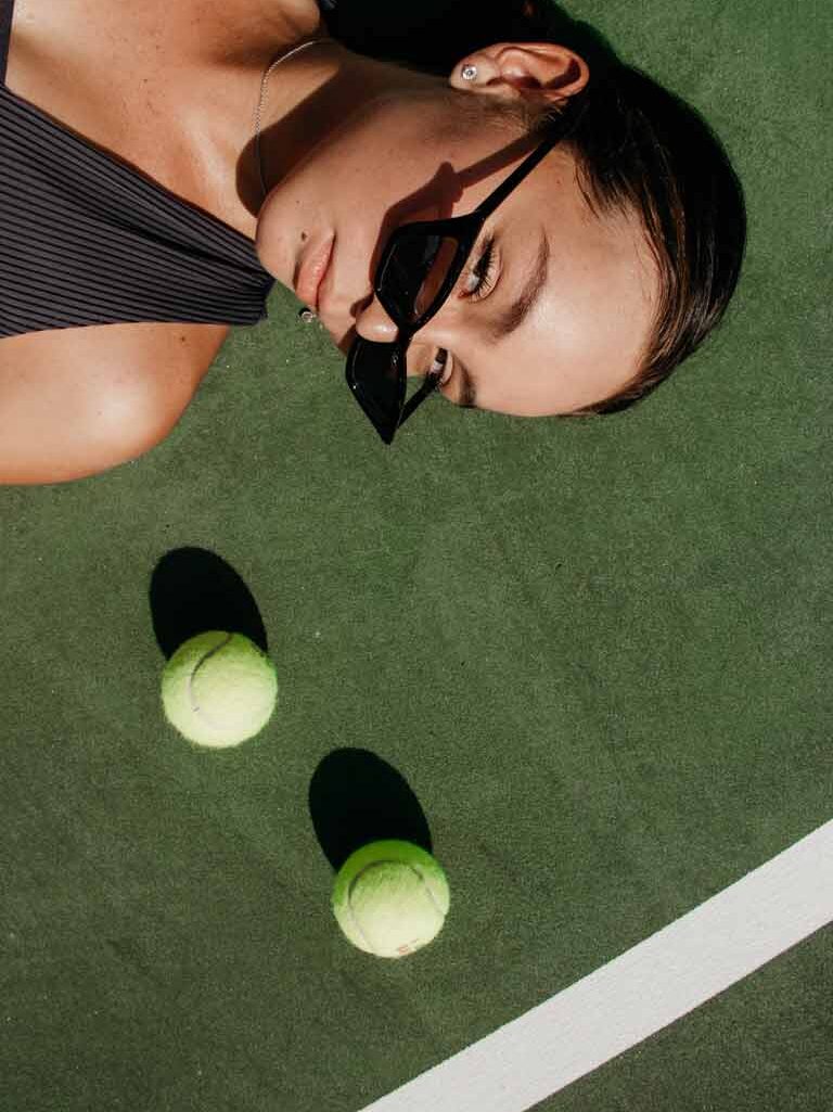 A cropped woman wearing black sunglasses is lying beside two tennis balls on a tennis court.