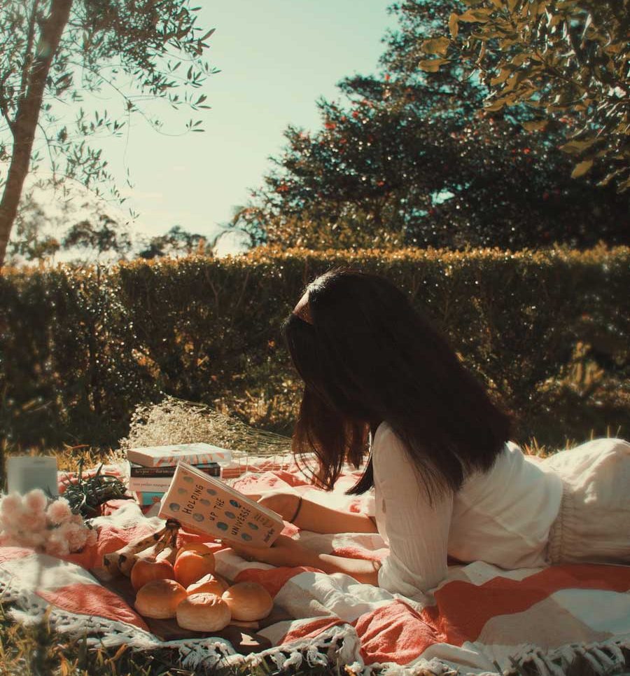 a woman lying down on a picnic blanket enjoying her time reading a book in a peaceful park